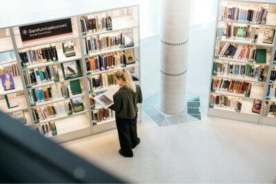 Ung kvinne står med ryggen til blant reoler fulle av bøker på et bibliotek.