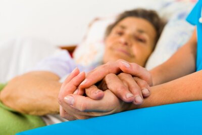 Bilde av en eldre dame som ligger i sengen mens en annen person holder hendene.