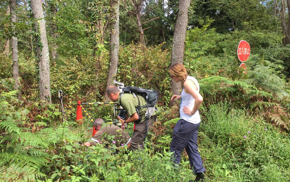 en kvinne og to menn driver med feltarbeid i en skog , der har ulike måleapparat med seg, og vi ser et japansk skilt i bakgrunnen