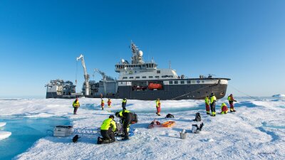 Forskningsskipet Kronprins Haakon ligger i isen. I front står en mengde forskere på isen i gule og røde klør og tar prøver