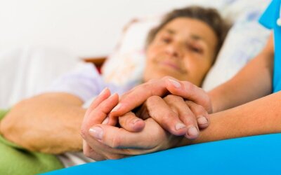 Bilde som viser en pleier som holder hendene til en pasient som ligger i sengen.