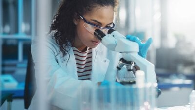 Ung jente med mørkt, krøllet hår ser ned i et mikroskop. Hun har på seg labfrakk over en stripete t-skjorte