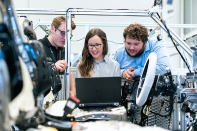 Tre unge ingeniører ser på en laptop i et produksjonslokale for biler